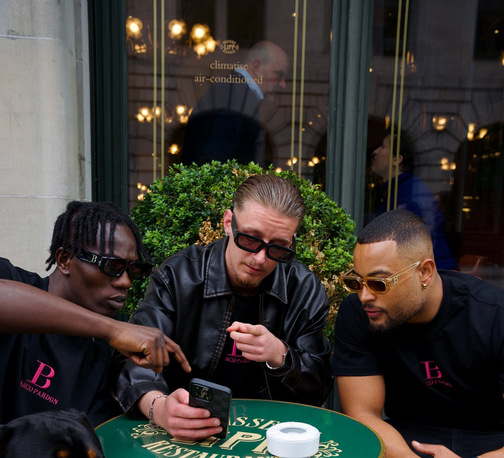 Three men in black BLANCO PARDON T-shirts and sunglasses at a cafe table