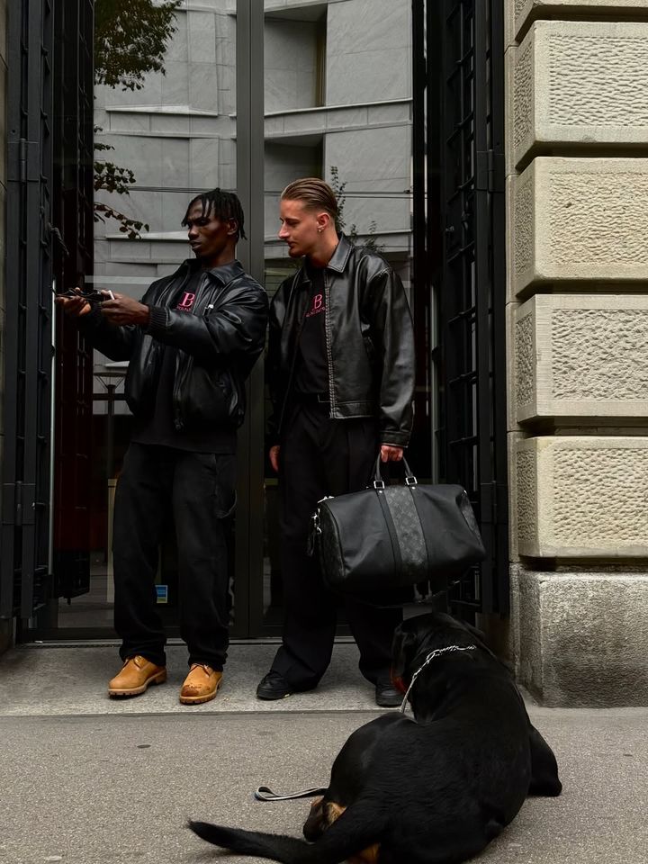 Two men in black BLANCO PARDON outfits with a Rottweiler at a doorway